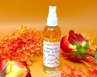 May include: A clear glass bottle with a white spray nozzle and a white label that reads "Eucalyptus Blooms Tangerine Shower & Room Spray". The bottle is sitting on a brown surface with orange and red flowers.