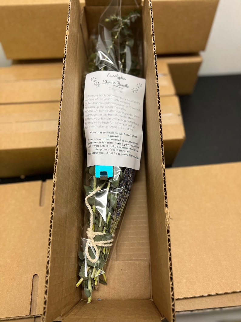 May include: A bundle of eucalyptus and lavender tied with twine in a clear plastic bag. The bundle is inside a cardboard box. The text on the label reads "Eucalyptus Shower Bundle Directions: Remove hook before placing on a dry surface where your bundle will hang. Run bundle under hot water for 10 seconds. Warm up the oils in the stems. Squeeze bundle after running under water. Hang your bundle by the loop on the hook. Enjoy the oils from inside the stems. Enjoy while fresh for 1-2 weeks and then throw away after as for or once it dries. Note that some pieces will fall off after you see a white powder-like substance on the leaves. It is normal during growth and not mold. If you detect mold, discard immediately. Keep out of reach from pets. Product should not be consumed internally."
