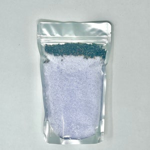 May include: A clear plastic pouch filled with lavender bath salts. The salts are a light purple color and there are dried lavender buds on top.