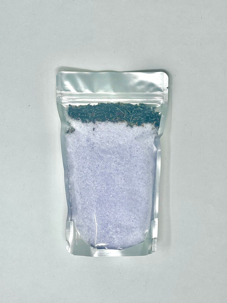 May include: A clear plastic pouch filled with lavender bath salts. The salts are a light purple color and there are dried lavender buds on top.