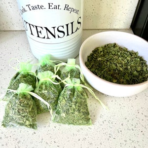 May include: Six small green mesh bags filled with dried herbs are arranged on a white countertop. A white bowl filled with dried herbs sits to the right. A white utensil holder with the words "Cook. Taste. Eat. Repeat. UTENSILS" is in the background.