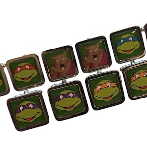 May include: A collection of square metal cufflinks featuring colorful graphic prints of Teenage Mutant Ninja Turtles characters, including Leonardo, Michelangelo, Donatello, Raphael, and Master Splinter, set against a green background.