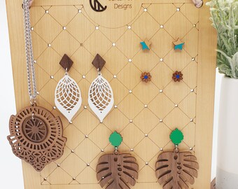 May include: Wooden jewelry display stand with a grid pattern. The stand features a variety of wooden earrings, including leaf-shaped earrings, star-shaped earrings, and monstera leaf earrings. The earrings are painted in various colors, including green, blue, and brown. The stand also features a wooden necklace with a circular design.