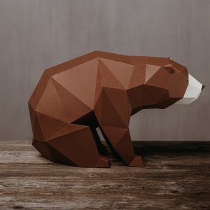 Può includere: Una scultura di orso di carta marrone con accenti bianchi sul viso. L'orso è seduto su una superficie di legno.
