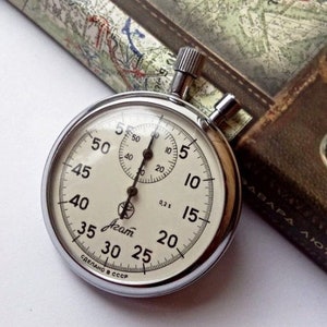 May include: A silver stopwatch with a white face and black markings. The stopwatch has a large dial with numbers from 5 to 60, and a smaller dial with numbers from 0 to 30. The stopwatch is made in the USSR and has the word "Акам" printed on the face.