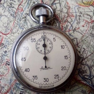 May include: A silver pocket watch with a white face and black hands. The watch has a black second hand and a small dial with black numbers. The watch face has the word "Azam" printed on it and the text "СДЕЛАНО В СССР" printed on the bottom.