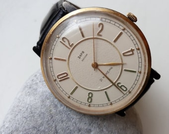 May include: A gold-tone wristwatch with a white face and black leather band. The watch face has black Roman numeral hour markers and a black second hand. The watch face also has the text "ЗАРЯ" and "21 К МЕХ" in Cyrillic.