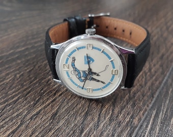 May include: A silver wristwatch with a black leather band. The watch face features a white background with a blue and white illustration of a hockey player. The watch face also has black Roman numeral hour markers.