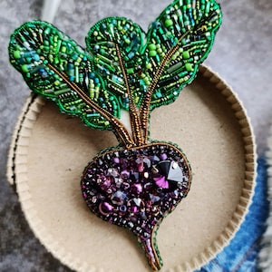 Könnte beinhalten: Eine handgefertigte Rote-Bete-Brosche mit einer tiefvioletten Wurzel, die mit facettierten Kristallen und Perlen verziert ist, gekrönt von leuchtend grünen Perlenblättern. Die aufwendige Stickerei verwendet verschiedene Grüntöne aus Glasperlen.