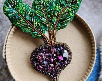 May include: A handcrafted beet brooch featuring a deep purple root embellished with faceted crystals and beads, topped with vibrant green beaded leaves. The intricate embroidery uses various shades of green glass beads and metallic thread to define the foliage.