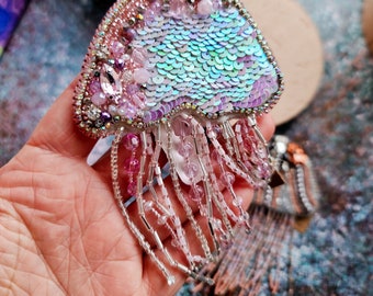 Puede incluir: Un broche de medusa de cuentas rosadas e iridiscentes con una franja de cuentas plateadas y rosadas.