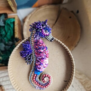 Puede incluir: Un broche de caballito de mar de cuentas moradas y rosas con lentejuelas iridiscentes. El caballito de mar est&aacute; enroscado en espiral con un gran cristal en el centro.