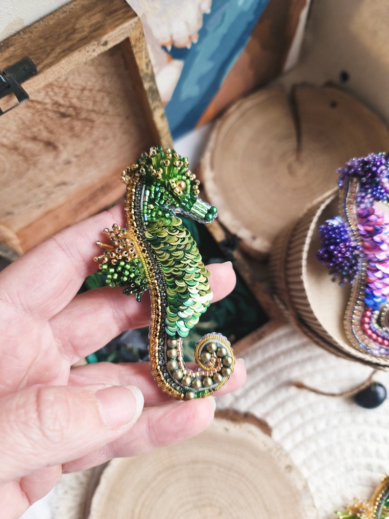 Puede incluir: Un broche de caballito de mar de cuentas verdes y doradas. El caballito de mar est&aacute; hecho de cuentas verdes y doradas y tiene un dise&ntilde;o detallado.