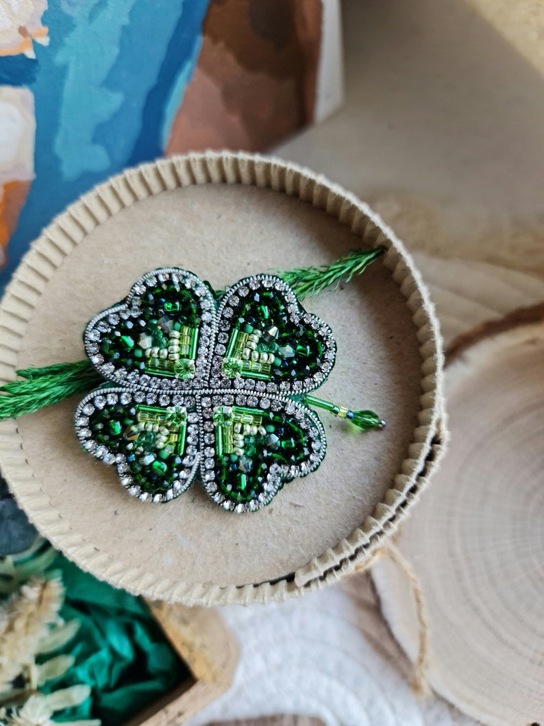 K&ouml;nnte beinhalten: Eine gr&uuml;ne vierbl&auml;ttrige Kleeblatt-Brosche aus Perlen und Strasssteinen. Die Brosche wird auf einem braunen Karton pr&auml;sentiert.