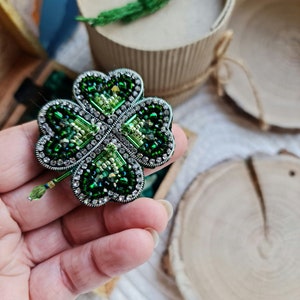 K&ouml;nnte beinhalten: Eine gr&uuml;ne vierbl&auml;ttrige Kleeblatt-Brosche mit silbernen Akzenten und Strasssteinen. Die Brosche ist aus Perlen gefertigt und ein handgemachtes Schmuckst&uuml;ck.