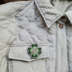 K&ouml;nnte beinhalten: Eine gr&uuml;ne vierbl&auml;ttrige Kleeblatt-Brosche mit Strasssteinen, die an einer hellgrauen gesteppten Jacke mit einer Tasche befestigt ist. Die Jacke hat ein Etikett mit der Aufschrift "DAISY STREET".