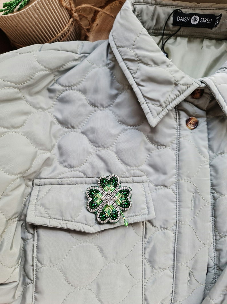 K&ouml;nnte beinhalten: Eine gr&uuml;ne vierbl&auml;ttrige Kleeblatt-Brosche mit Strasssteinen, die an einer hellgrauen gesteppten Jacke mit einer Tasche befestigt ist. Die Jacke hat ein Etikett mit der Aufschrift "DAISY STREET".
