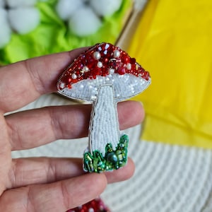Puede incluir: Un broche de hongo rojo y blanco con hojas verdes de cuentas. El hongo tiene un tallo blanco y una tapa roja con cuentas blancas alrededor del borde.