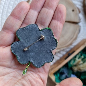 K&ouml;nnte beinhalten: Eine gr&uuml;ne vierbl&auml;ttrige Kleeblatt-Brosche mit einer silbernen Nadel auf der R&uuml;ckseite. Die Brosche ist aus Stoff und Perlen gefertigt.