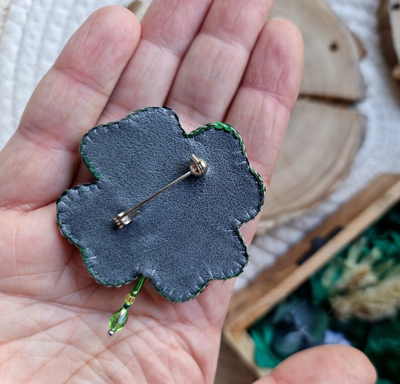 K&ouml;nnte beinhalten: Eine gr&uuml;ne vierbl&auml;ttrige Kleeblatt-Brosche mit einer silbernen Nadel auf der R&uuml;ckseite. Die Brosche ist aus Stoff und Perlen gefertigt.