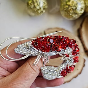May include: A red and silver beaded shrimp brooch. The shrimp is made of red and silver beads and sequins. The shrimp is curled up in a realistic pose.