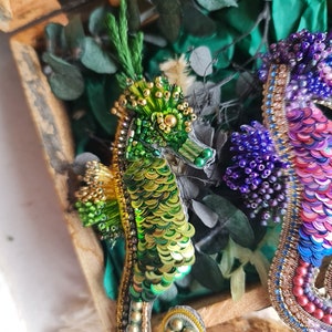 Puede incluir: Dos broches de caballito de mar hechos con cuentas, uno verde y dorado, el otro morado, rosa y azul, en una caja de madera con follaje verde.
