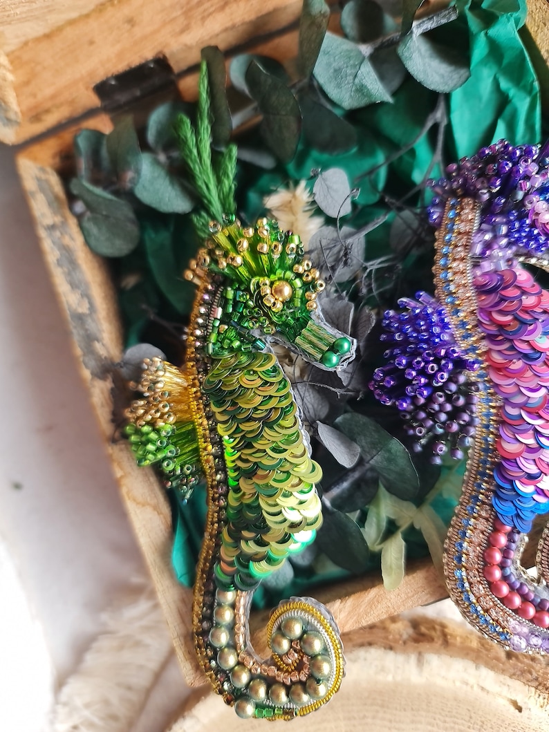 Puede incluir: Dos broches de caballito de mar hechos con cuentas, uno verde y dorado, el otro morado, rosa y azul, en una caja de madera con follaje verde.