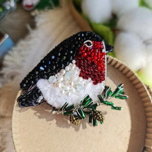 May include: A beaded robin brooch with red, white, and black accents. The robin is perched on a green beaded branch with a brown beaded pine cone.