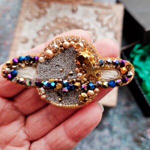 May include: A gold and silver beaded brooch with a star and a white crescent moon design. The brooch is shaped like a planet with a ring around it. The beads are iridescent and sparkle in the light.