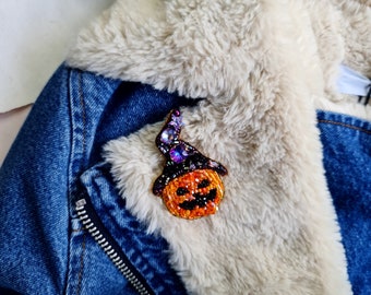 Może przedstawiać: Brozka w kształcie dyni z koralików z czarno-fioletowym kapeluszem czarownicy, broszka w stylu Halloween.