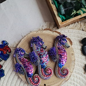 Puede incluir: Tres broches de caballito de mar hechos con cuentas en tonos de morado, rosa y azul. Los broches se muestran sobre una superficie de madera.