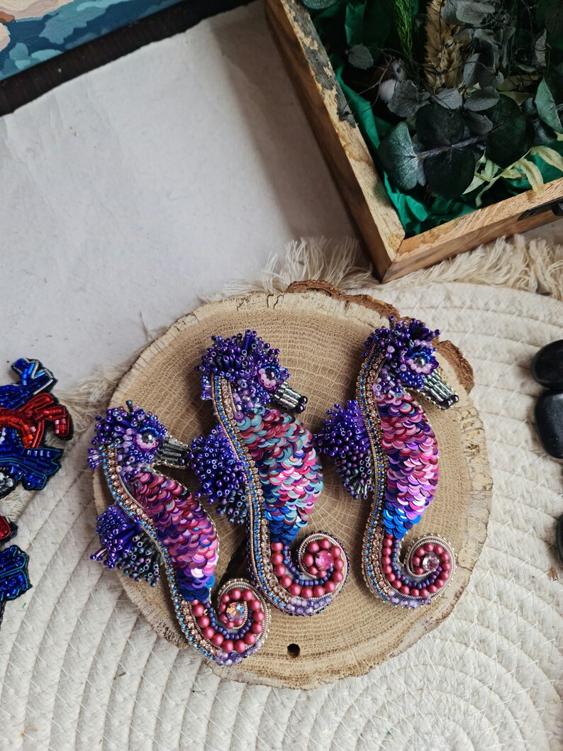 Puede incluir: Tres broches de caballito de mar hechos con cuentas en tonos de morado, rosa y azul. Los broches se muestran sobre una superficie de madera.