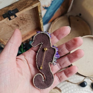 Puede incluir: Un broche de caballito de mar de cuentas moradas y plateadas con un alfiler dorado en la parte posterior. El broche est&aacute; sobre un respaldo de fieltro marr&oacute;n.