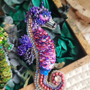 Puede incluir: Un broche de caballito de mar con cuentas y lentejuelas iridiscentes de color rosa, morado y azul. El caballito de mar est&aacute; enroscado en espiral con un gran cristal en la cola.