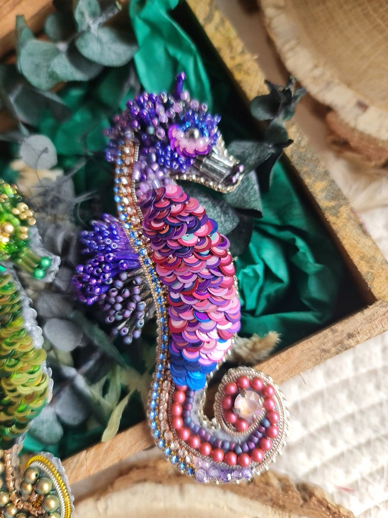 Puede incluir: Un broche de caballito de mar con cuentas y lentejuelas iridiscentes de color rosa, morado y azul. El caballito de mar est&aacute; enroscado en espiral con un gran cristal en la cola.