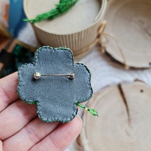 K&ouml;nnte beinhalten: Eine gr&uuml;ne Kleeblatt-Brosche mit einem Perlenmuster und einer Stecknadel auf der R&uuml;ckseite. Die Brosche befindet sich auf einer Hand.
