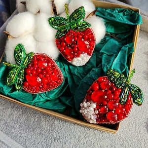 Puede incluir: Tres broches de fresa de cuentas en una caja de regalo. Los broches están hechos con cuentas rojas, verdes y blancas. Los broches están en una caja forrada con papel de seda verde.