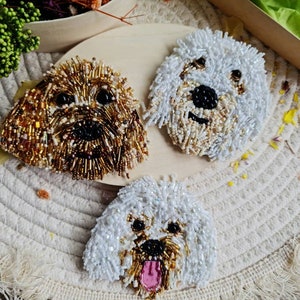 Puede incluir: Tres broches de perro de cuentas, uno marrón y dorado, uno blanco y uno blanco con la lengua rosa. Los broches están sobre un fondo blanco con flecos.