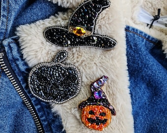 Może przedstawiać: Trzy broszki w stylu Halloween: czarny kapelusz czarownicy, czarna dynia i pomarańczowa dynia z fioletowym kapeluszem, są przypięte do dżinsowej kurtki.
