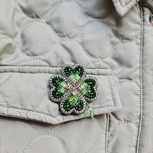 K&ouml;nnte beinhalten: Eine gr&uuml;ne vierbl&auml;ttrige Kleeblatt-Brosche mit gr&uuml;nen und klaren Strasssteinen. Die Brosche befindet sich auf einer grauen gesteppten Jacke.