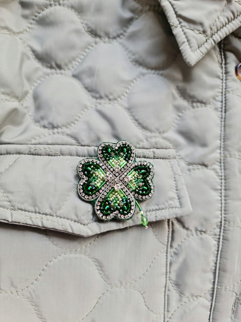 K&ouml;nnte beinhalten: Eine gr&uuml;ne vierbl&auml;ttrige Kleeblatt-Brosche mit gr&uuml;nen und klaren Strasssteinen. Die Brosche befindet sich auf einer grauen gesteppten Jacke.