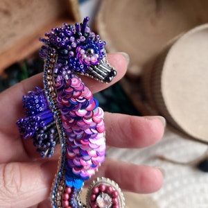 Puede incluir: Un broche de caballito de mar con cuentas y lentejuelas iridiscentes de color rosa, morado y azul. El caballito de mar tiene una cabeza detallada con una corona y un cristal en su cola.