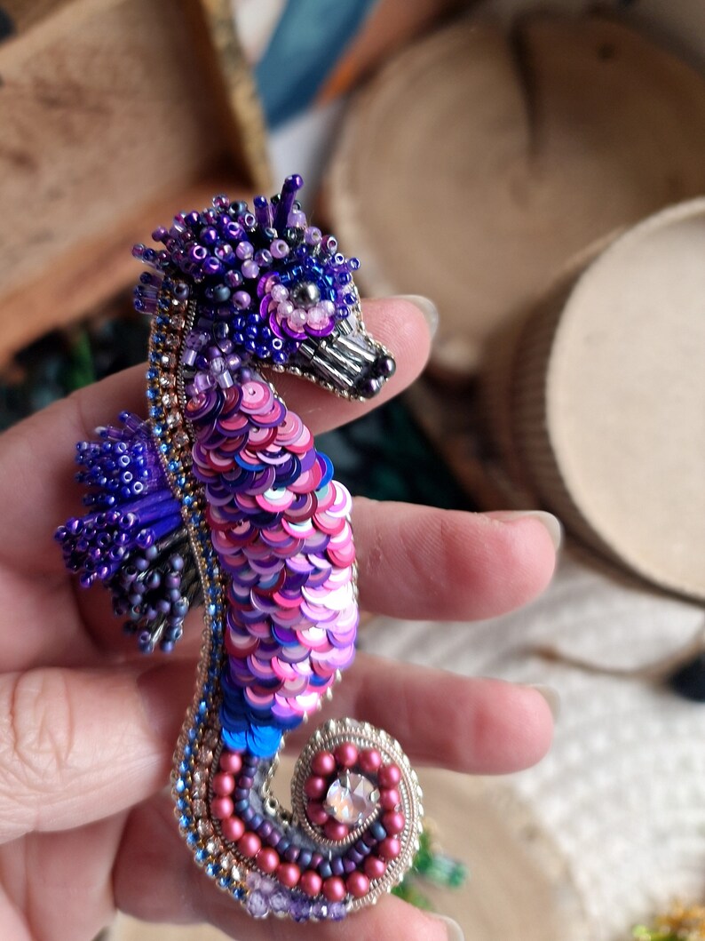 Puede incluir: Un broche de caballito de mar con cuentas y lentejuelas iridiscentes de color rosa, morado y azul. El caballito de mar tiene una cabeza detallada con una corona y un cristal en su cola.