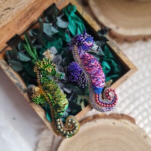 Puede incluir: Dos broches de caballito de mar hechos con cuentas, uno verde y otro morado, en una caja de madera con follaje verde.