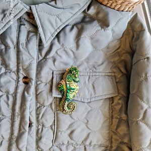 Puede incluir: Un broche de caballito de mar de cuentas verdes y doradas prendido en una chaqueta acolchada gris.
