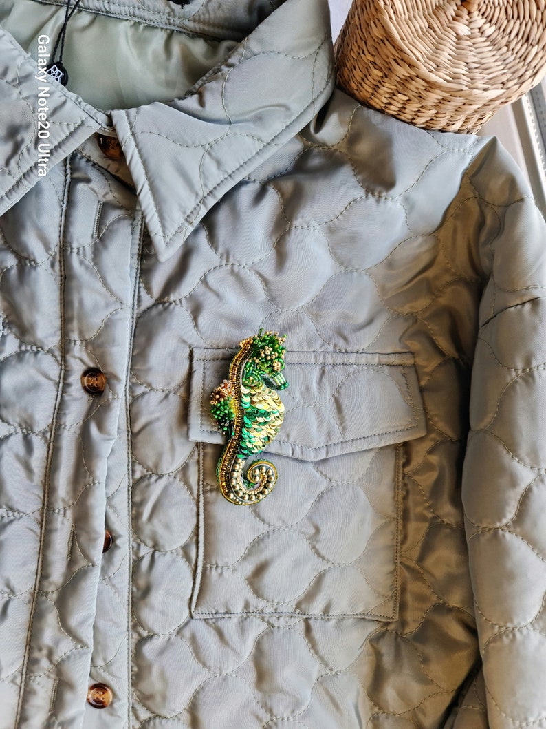 Puede incluir: Un broche de caballito de mar de cuentas verdes y doradas prendido en una chaqueta acolchada gris.