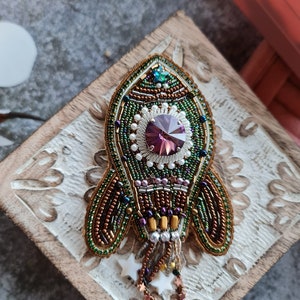 May include: A green, brown, and gold beaded rocket ship brooch with a purple rhinestone in the center. The brooch is on a wooden surface.