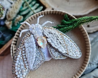 May include: A white beaded moth brooch with iridescent accents. The moth has delicate wings and a detailed body. The brooch is displayed on a white background.