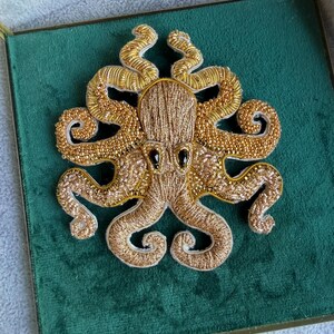 Puede incluir: Un broche de pulpo en tono dorado elaborado con intrincados bordados y abalorios. El diseño presenta un cuerpo central texturizado, ocho tentáculos rizados adornados con cuentas doradas metálicas y dos ojos oscuros en forma de lágrima sobre fondo blanco.