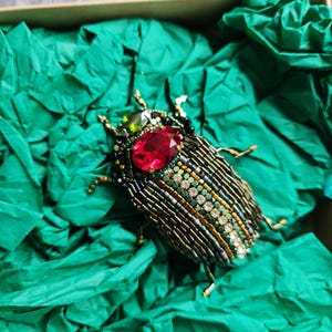 May include: A beaded insect brooch with a large red gemstone and smaller green gemstone accents. The body is detailed with gold, green, and black beads. The brooch is displayed on green tissue paper inside a box.